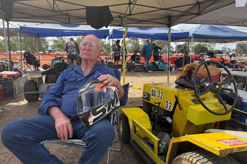 A man with his yellow race mower, cradling a helmet.