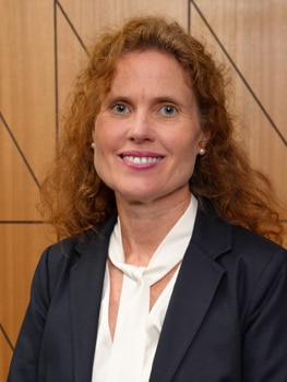 A woman with long curly red hair in a blue blazer and white silky top sits in front of a wood panel wall, smiling.
