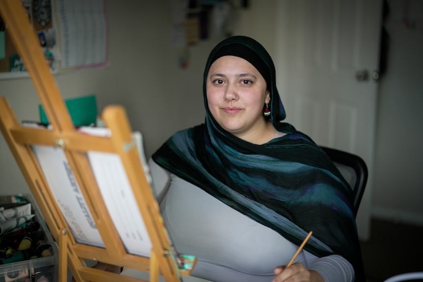 A woman sits at a painting easle looking at camera.