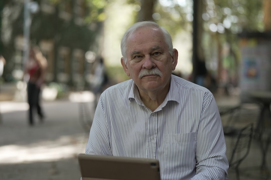 A man with white hair and moustache sits outside looking seriously into camera.