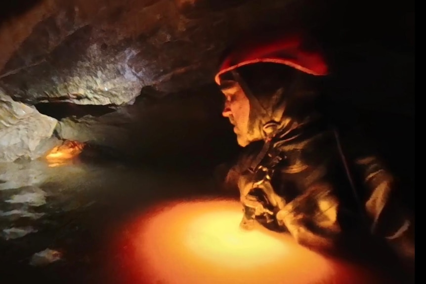 A man in a full body wet suit and orange helmet wading through chest-deep water in a dark cave.