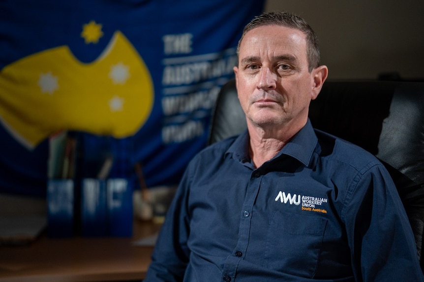 A man in a blue Australian Workers' Union shirt with the union flag on the wall behind him.