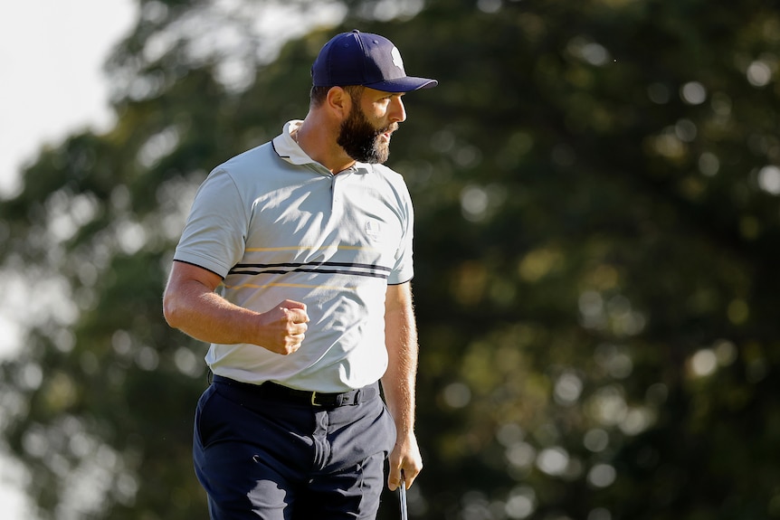 A bearded man wearing a blue cap and white golf shirt clenches his fist in celebration.