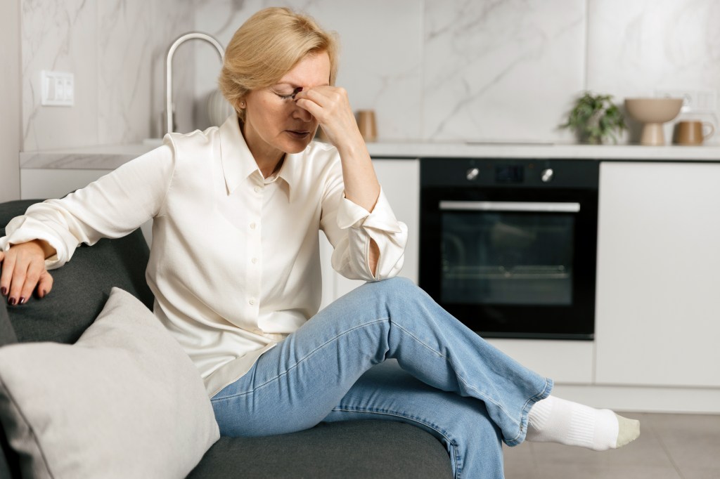 A middle-aged woman sits on a modern grey couch, holding her temples due to a headache.