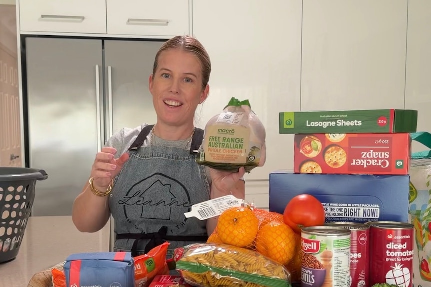A woman in an apron holding a chicken near a kitchen bench full of groceries
