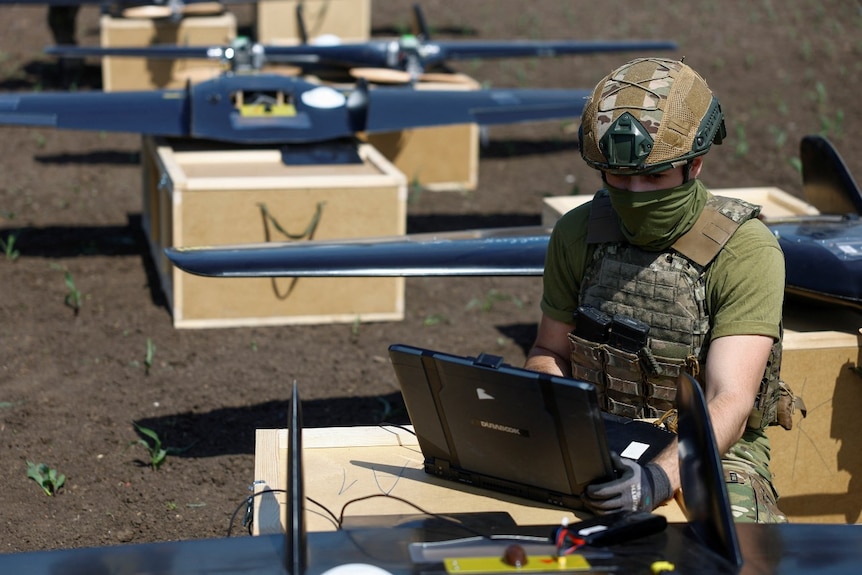 A soldier sitting outdoors and looking at a laptop. The soldier is surrounded by drones.