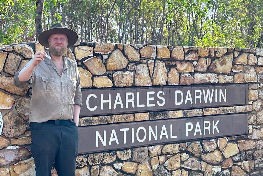 An ecologist with a beard wears a wide-brimmed hat as he stands in a national park and holds up a bug.