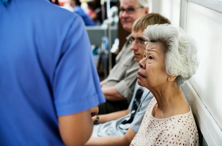 People seated in a waiting area and a person standing in blue scrubs seen from behind