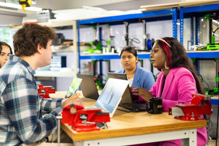 Several people including a Black woman in fuscia blazer, South Asian woman in lavender blazer and white man in checked flannel plaid stand gathered around high tables with laptops and what appear to be red vice grips on tables in front of utility shelving with varied equipment.