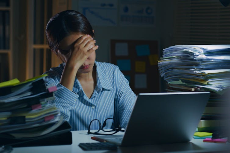 A woman rests her forehead against her hand while sitting at a desk piled high with paperwork