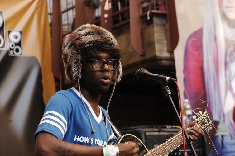 Lightspeed Champion playing guitar in a blue shirt and furry trapper hat.