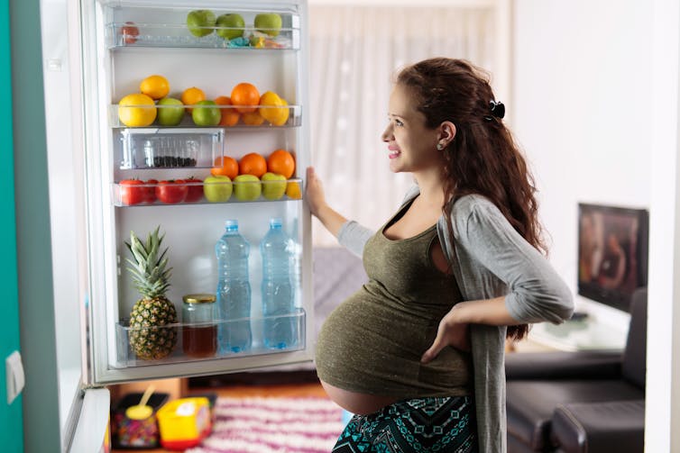 Heavily pregnant woman stands in front of open fridge, which is stocked with healthy food on display