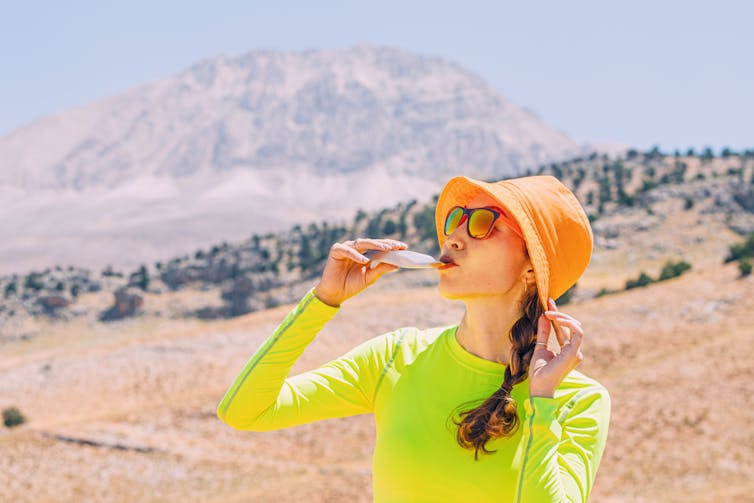 A woman wearing a hat and athletic gear slurps down an energy gel on a sunny day in the desert.