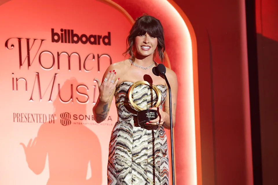 Ella Langley holds a Billboard Women in Music award while speaking at a microphone onstage, with event signage behind her.