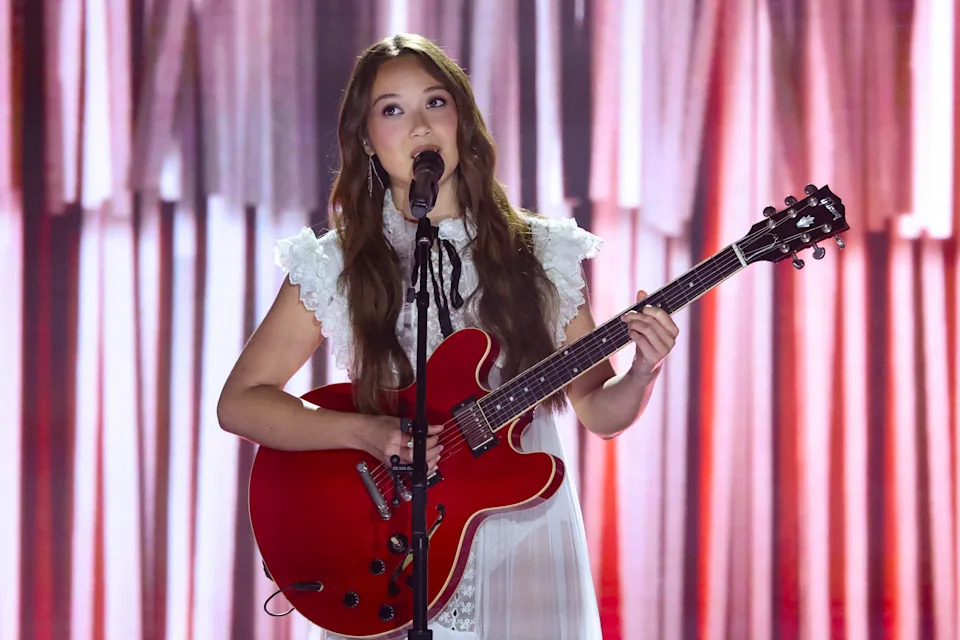 Laufey in a white dress sings into a microphone while playing a red electric guitar onstage with a softly lit backdrop.