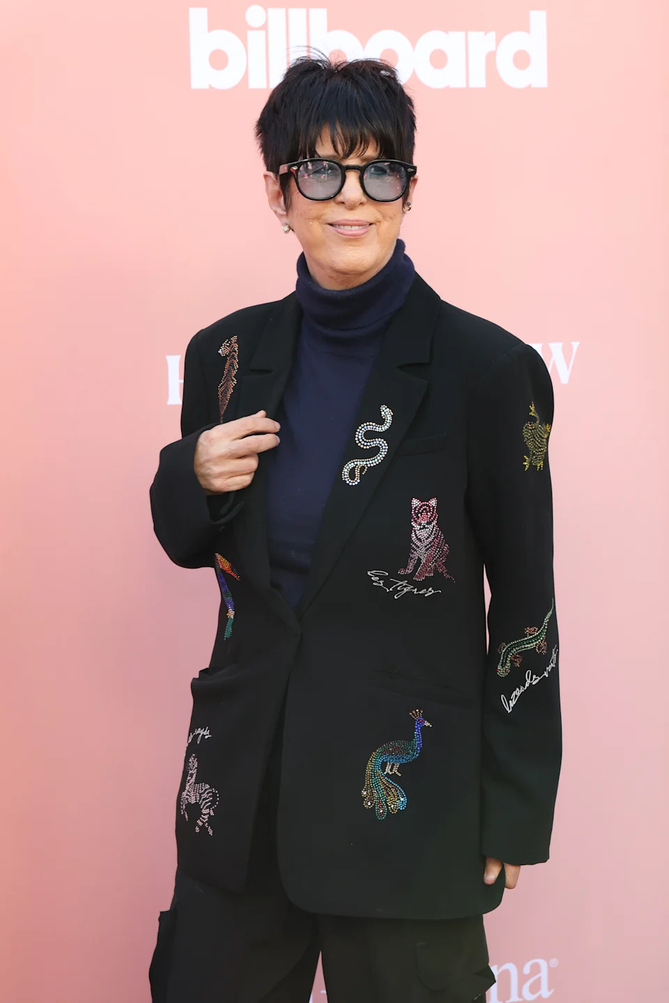 Diane Warren wearing black suit with embroidered animal designs and round sunglasses poses against a pink 'Billboard Women in Music' backdrop.