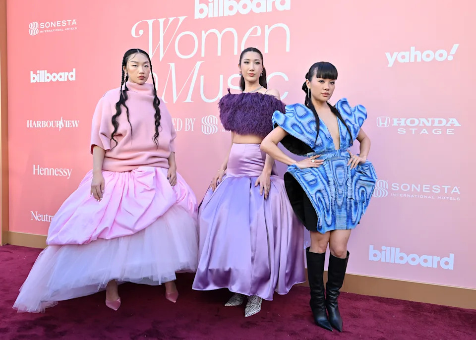 Audrey Nuna, Ejae and Rei Ami in avant-garde pink, purple, and blue dresses pose on a red carpet in front of a 'Billboard Women in Music' backdrop.