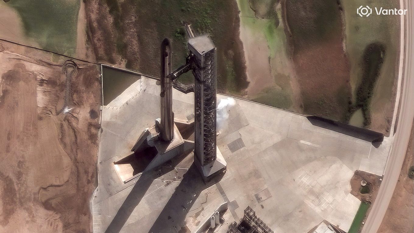 satellite photo of a black and silver rocket standing on a launch pad next to a large metallic tower
