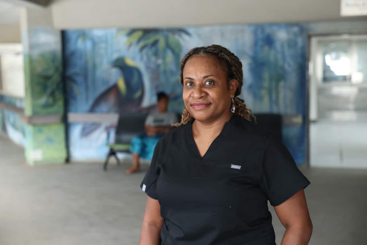 A woman with her hair pulled back and a slight smile stands in front of a painted mural in a hospital waiting area. She is wearing navy scrubs. 