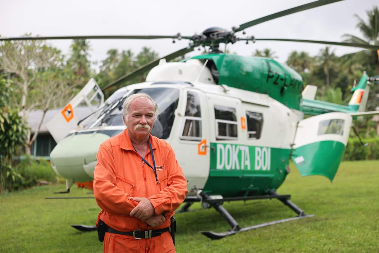 A man with a white mustache, wearing an orange jumpsuit, stands in front of a large green helicopter. Along the helicopter's side is text reading 'Dokta Boi'. 