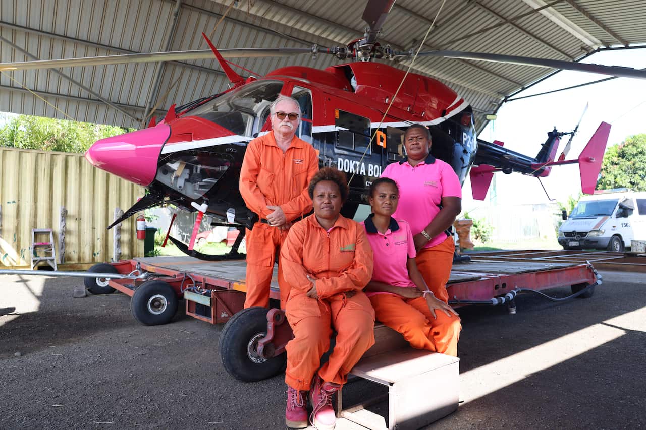A group of four people in bright pink shirts and orange jumpsuits stand in front of a red, blue and pink helicopter with the words 'Dokta Boi' on its side. 