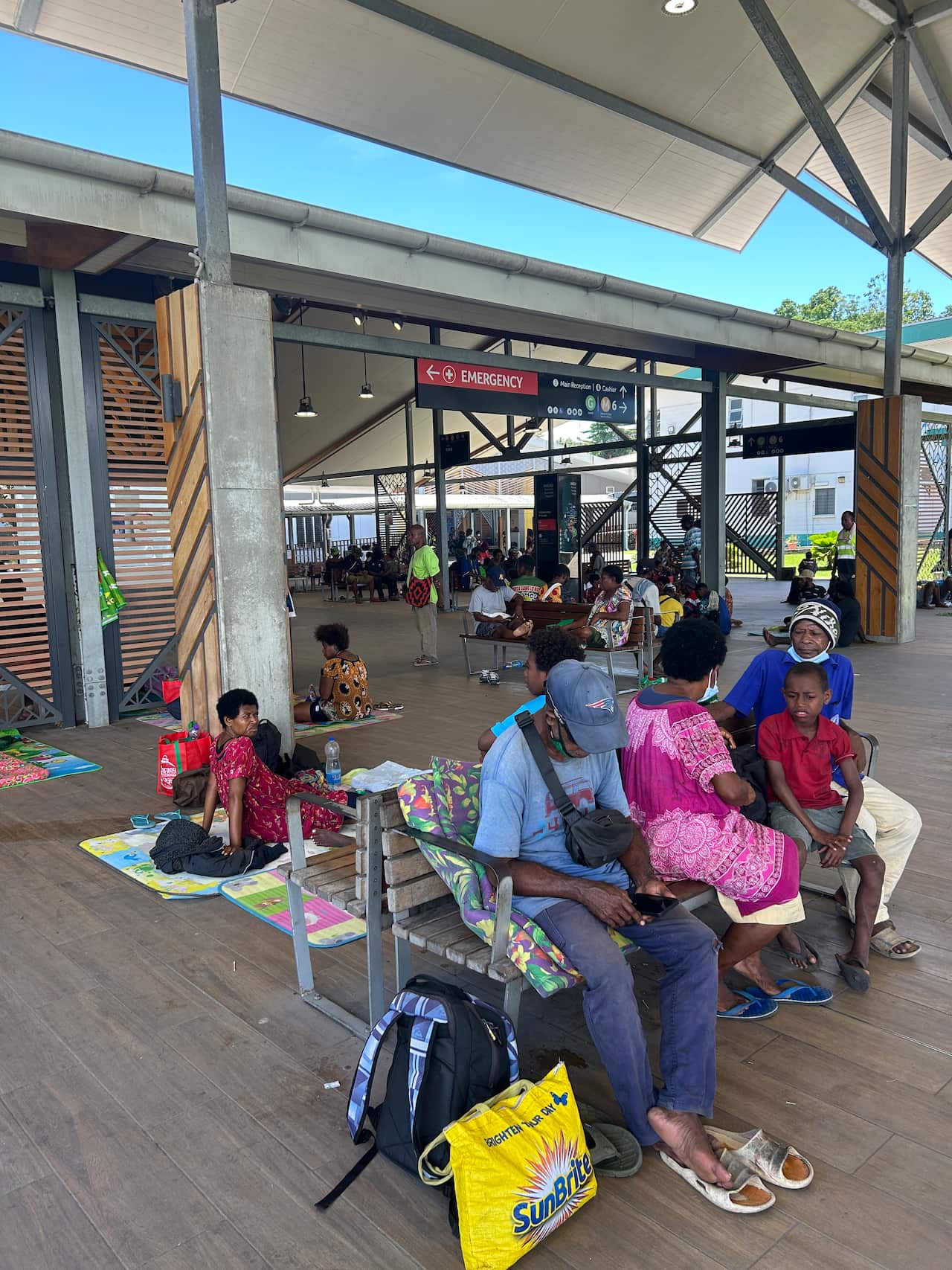 People are waiting outside the emergency department of a modern-looking hospital. Some are sitting on blankets on the floor, while others sit on wooden benches. 