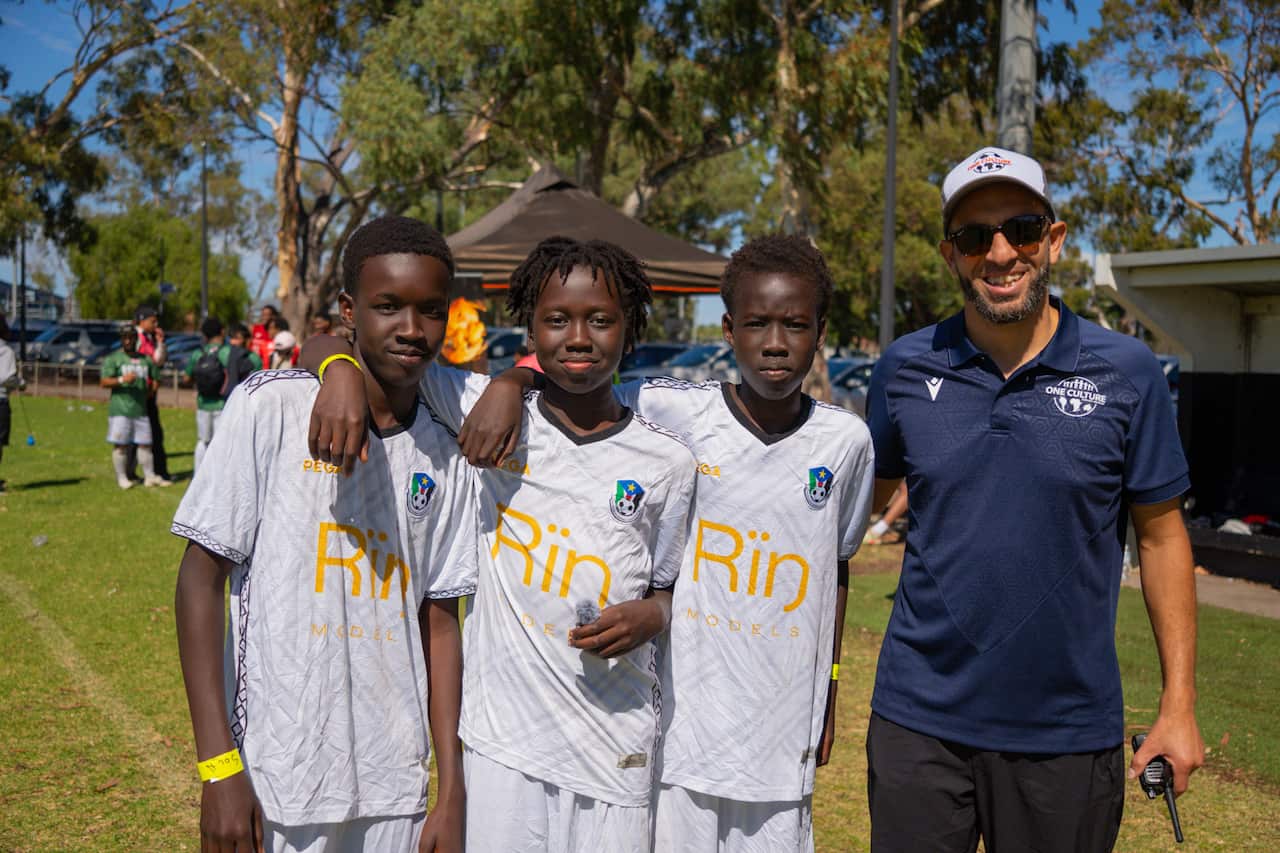 A man in a blue shirt stands next to three young football players in the field. They are wearing white football kits