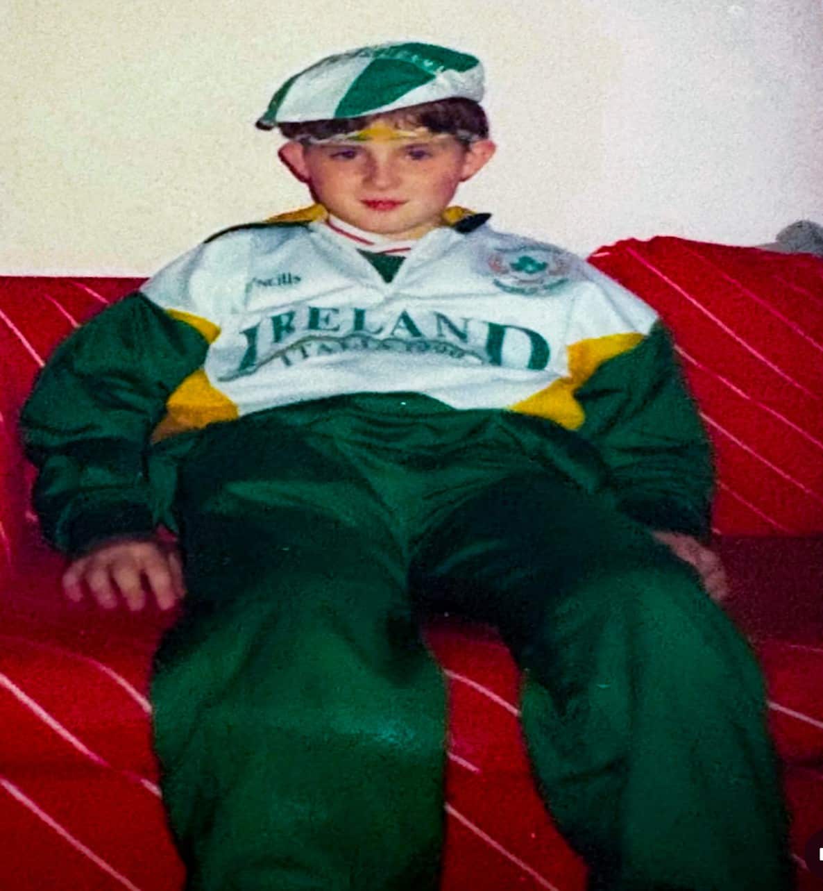 A young boy in a green-and-white Ireland football team tracksuit and matching cap is sitting on a red couch.