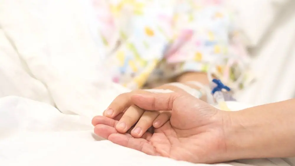 A close-up of an adult gently holding a child’s hand in a hospital bed. The child is wearing a colorful hospital gown and has a medical IV line attached, conveying comfort, care, and support during treatment.