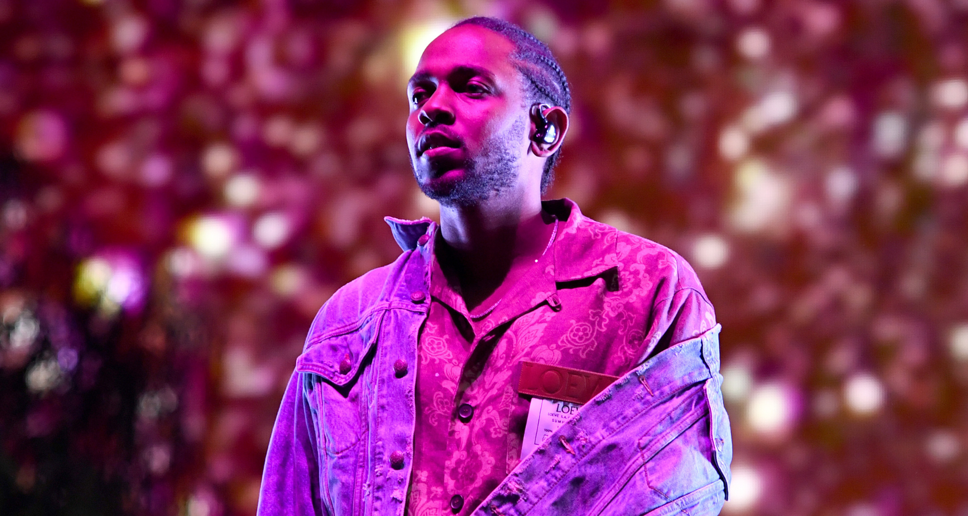 Kendrick Lamar performing on stage, wearing a patterned shirt and denim jacket, with a colorful, blurred background.