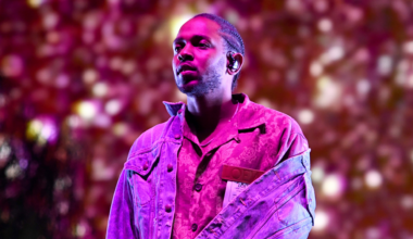 Kendrick Lamar performing on stage, wearing a patterned shirt and denim jacket, with a colorful, blurred background.
