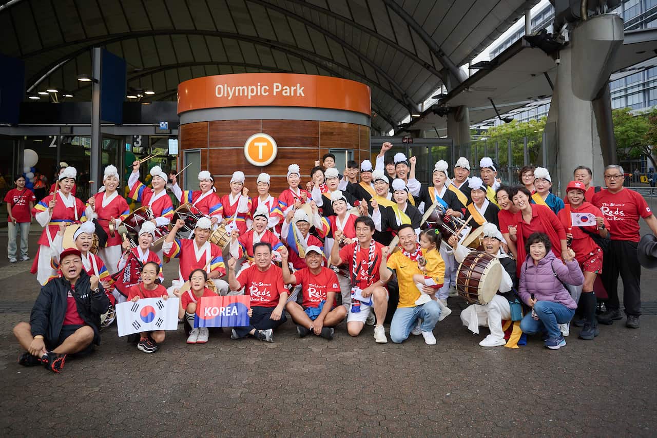 A group of football fans wearing red jerseys pose for a photo inside a train station. Some are holding drums and posters