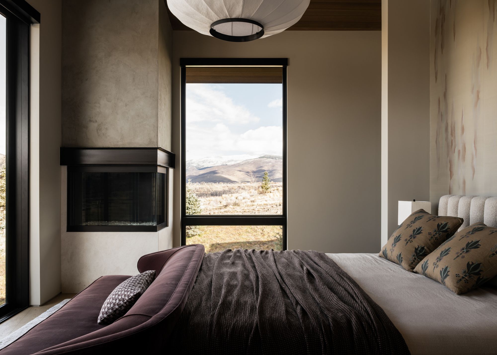 A neutral modern bedroom with a framed view of the mountainous surroundings and a purple sofa at the foot of the bed blending with the throw at the base of the bed below two throw pillows
