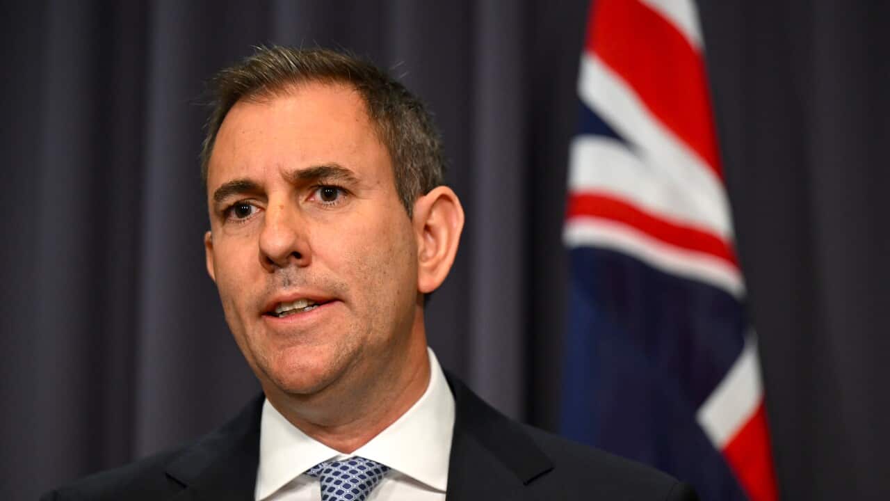 Treasurer Jim Chalmers stands in front of an Australian flag weaing a black suit and tie.