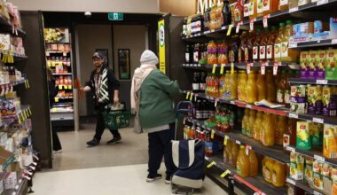 A person browses products at a supermarket aisle with another person seen with a shopping basket in the background
