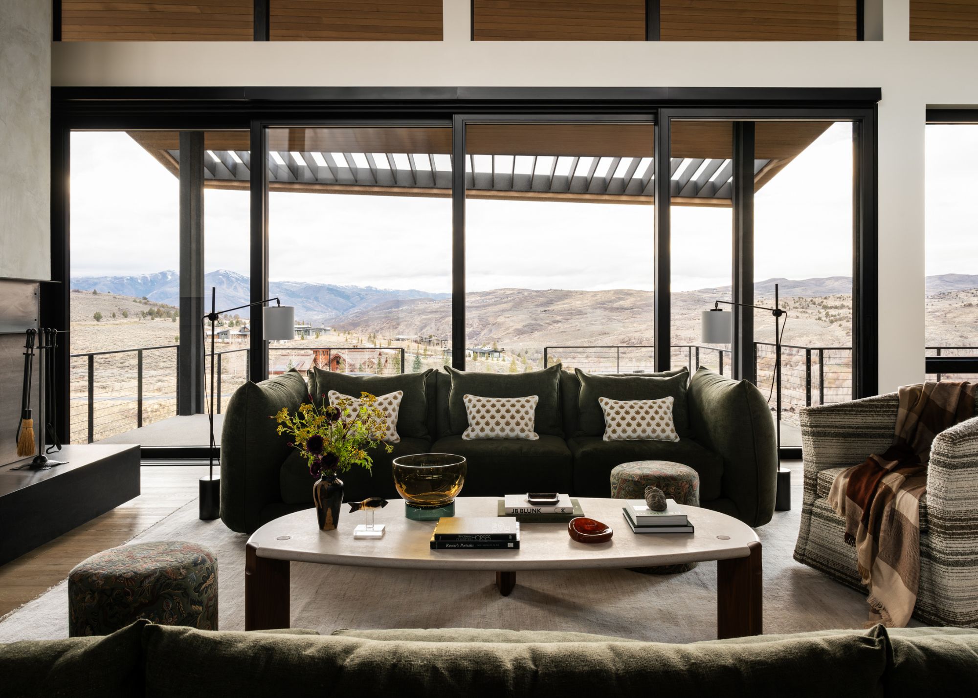A modern living space with black-framed floor-to-ceiling windows looking onto mountain views, with green sofas and a coffee table styled with decorative elements