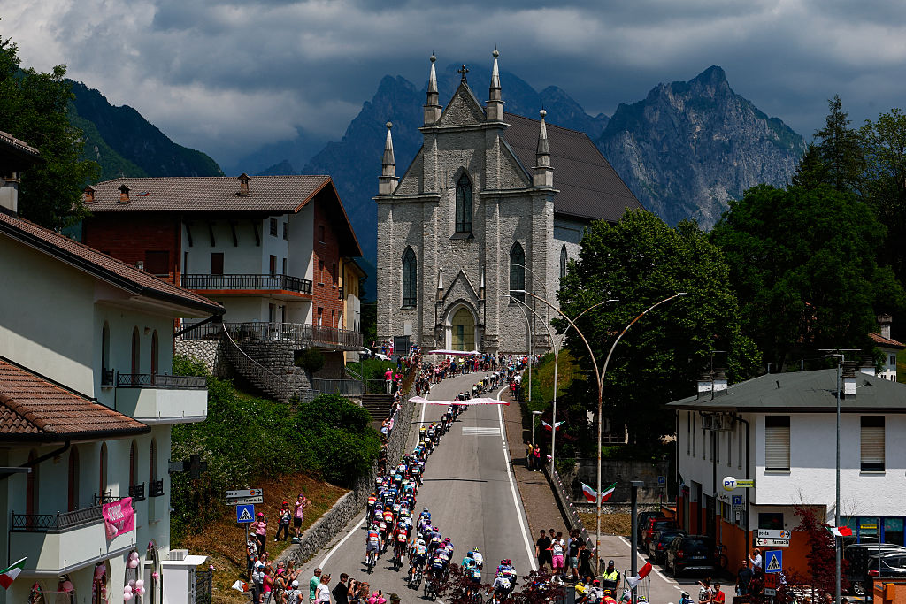 The pack of riders (peloton) cycle through the town of Sospirolo past the Santi Pietro e Paolo church near the Dolomite mountains during the 19th stage of the Giro d'Italia 2026 - Tour of Italy cycling race between Feltre and Alleghe, Italy, on May 29, 2026. (Photo by Luca Bettini / AFP)