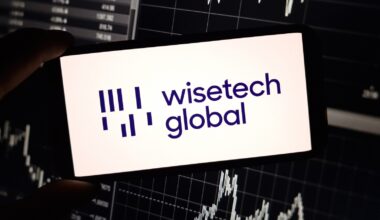 A close up of a smartphone displaying the WiseTech Global logo, in front of a board of stock prices