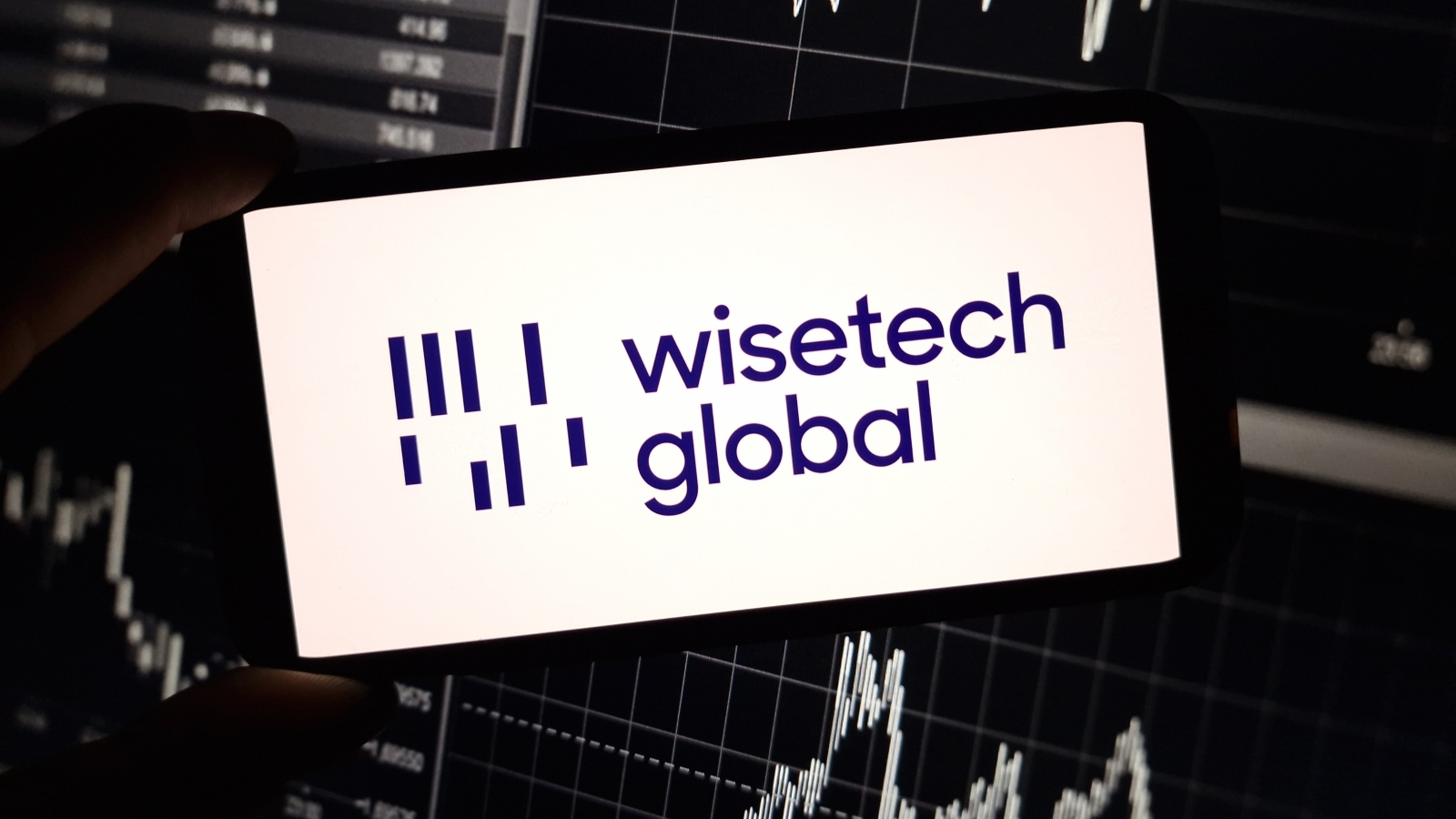 A close up of a smartphone displaying the WiseTech Global logo, in front of a board of stock prices