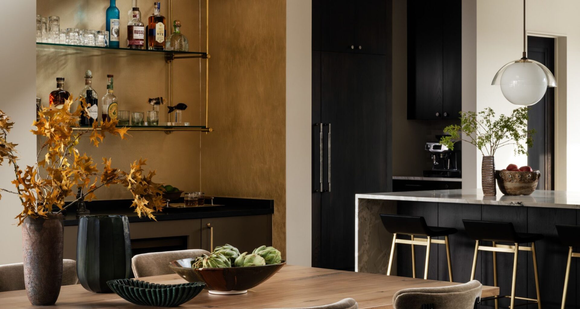 A modern dining space with a home bar in a niche with brass detailing and glass shelving next to the kitchen's island