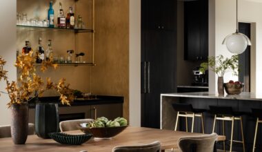 A modern dining space with a home bar in a niche with brass detailing and glass shelving next to the kitchen's island