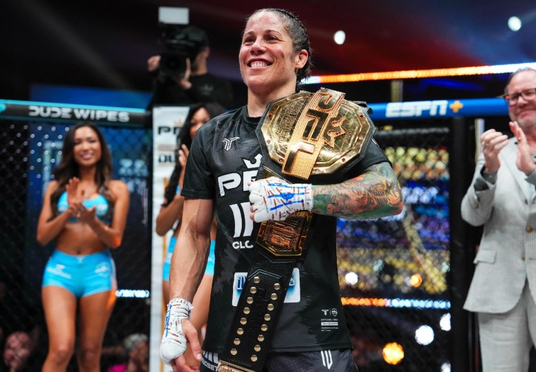 A woman stands in a ring gripping, with a heavily tattooed arm, a large metal gold toned belt over her left shoulder. A woman can be seen clapping over her shoulder.
