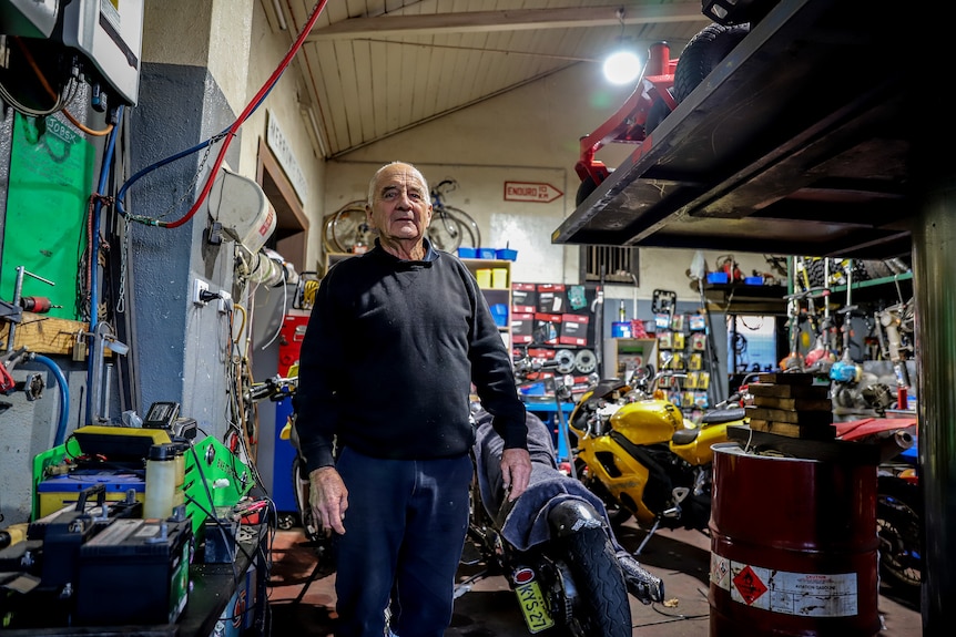 Older man wearing blue jumper stands in cluttered gar mechanic garage