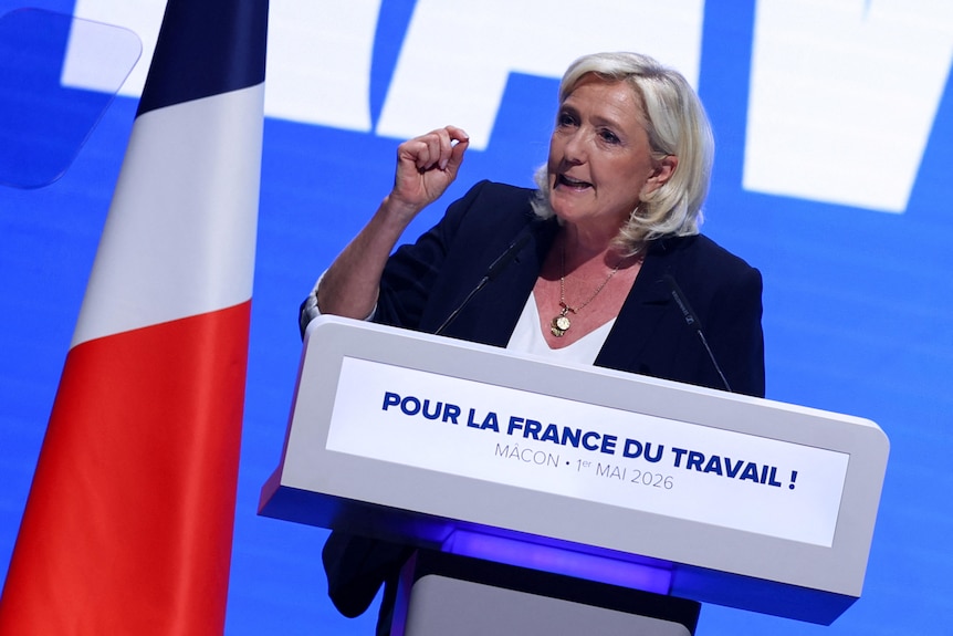 French far-right leader Marine Le Pen speaks behind a lectern near a French flag.