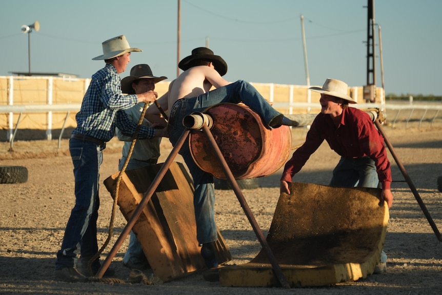 Young man in cowboy hat tries to jump over an obstacle as others help around him.