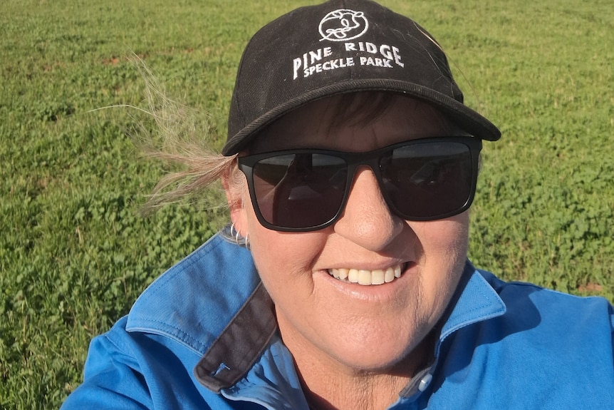 A women wearing black sunglasses and a cap takes a selfie on a green crop.