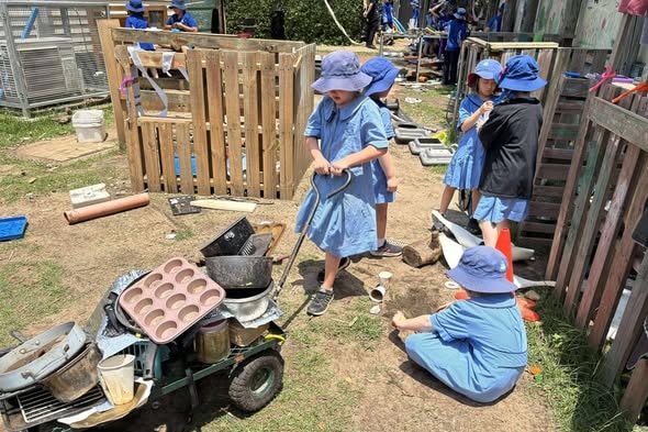 kids push a trolley full of containers and kitchen equipment through a messy yard with wooden crates around them
