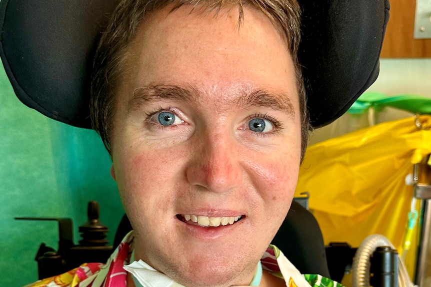 A smiling Caucasian man in a wheelchair with a tracheostomy tube