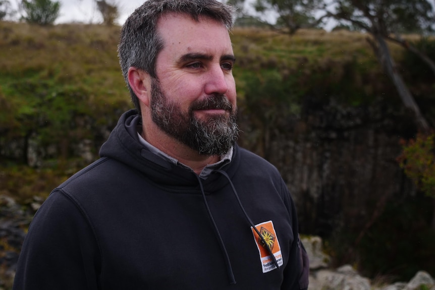 A man with a beard looks into the distance with greenery behind him