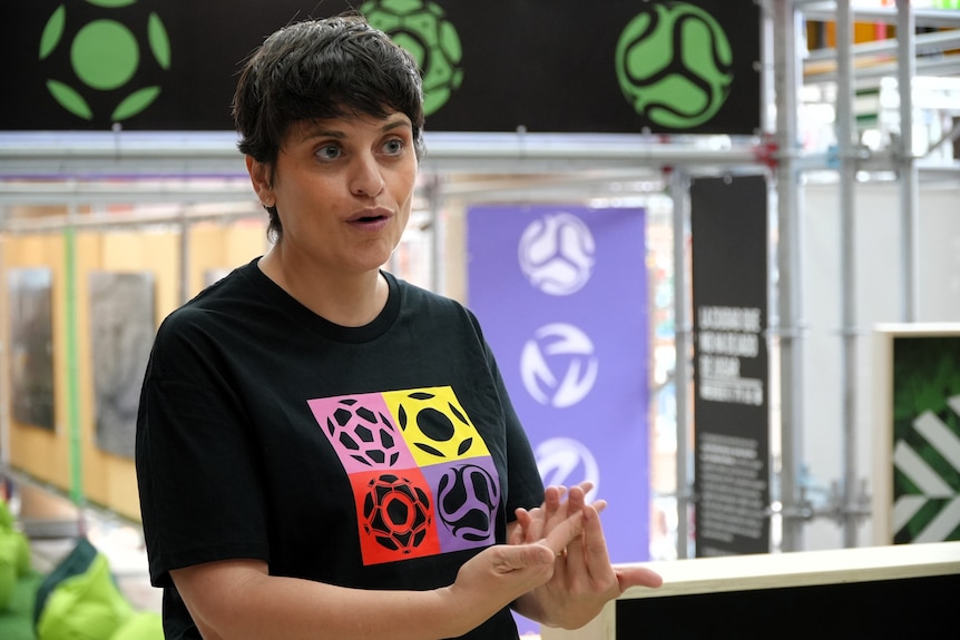 A woman with short brown hair wears a t-shirt with colourful soccer balls, and she speaks